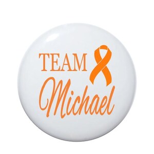 May include: White button with the text "TEAM Michael" in orange lettering and an orange ribbon.