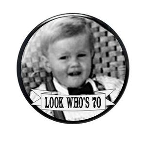 May include: A circular black and white button featuring a vintage photograph of a young child wearing a bow tie. Below the child, a white banner with black borders displays the text LOOK WHO'S 70 in a bold, stylized serif font.