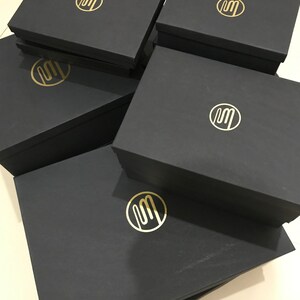 Puede incluir: Pila de cinco cajas de regalo negras con un logotipo dorado de la letra "M" en un c&iacute;rculo en cada caja.