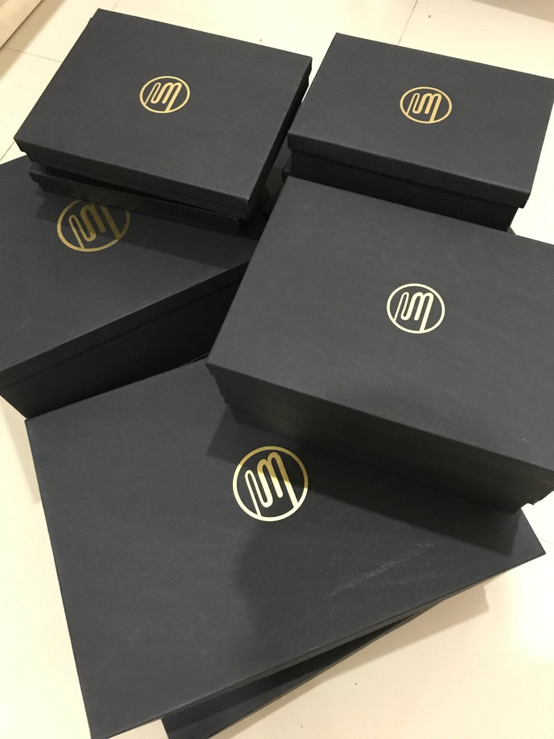 Puede incluir: Pila de cinco cajas de regalo negras con un logotipo dorado de la letra "M" en un c&iacute;rculo en cada caja.