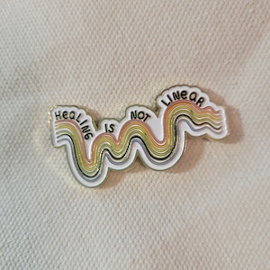 May include: A white enamel pin with the text "HEALING IS NOT LINEAR" in black lettering. The pin features a wavy design with rainbow-colored stripes. The pin has a gold border.