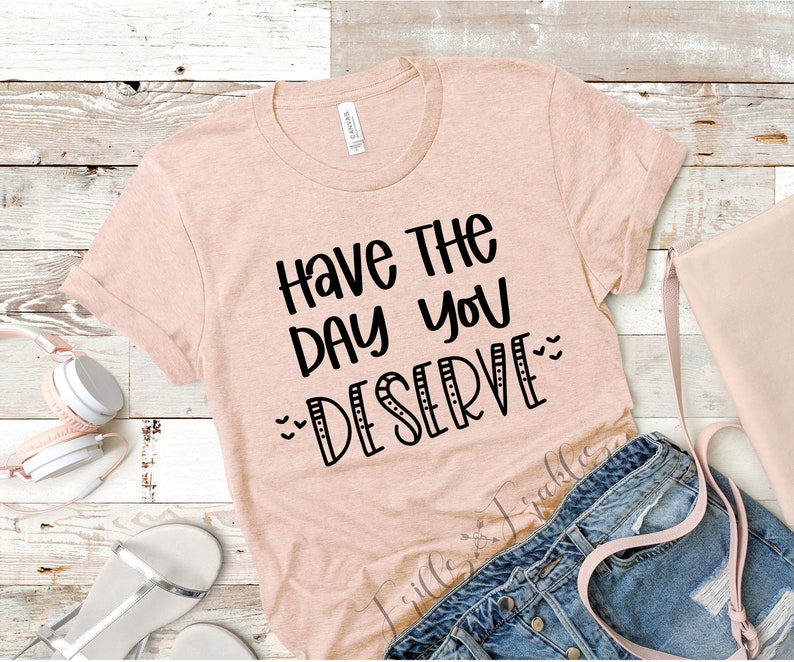 K&ouml;nnte beinhalten: Hellpfirsichfarbenes T-Shirt mit umgekrempelten &Auml;rmeln und dem schwarzen Schriftzug "Have The Day You Deserve". Das Shirt wird mit Jeansshorts, einer hellrosa Tasche und wei&szlig;en Kopfh&ouml;rern auf einer Holzoberfl&auml;che pr&auml;sentiert.