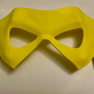 May include: A bright yellow mask with a distinctive shape, featuring eye openings and a curved design. The mask is made of a solid material and is likely for costume or cosplay purposes.