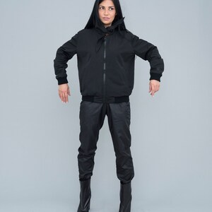 May include: A black bomber jacket with a high collar and a black zip. The jacket has a black drawstring at the waist and is worn with black trousers and black boots.