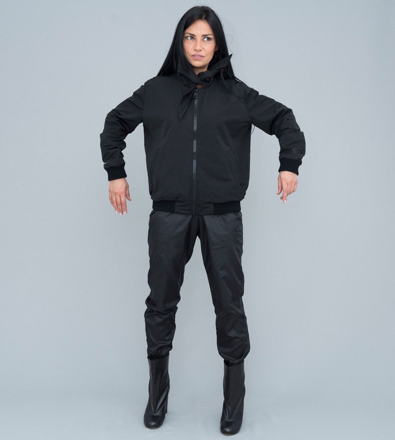 May include: A black bomber jacket with a high collar and a black zip. The jacket has a black drawstring at the waist and is worn with black trousers and black boots.