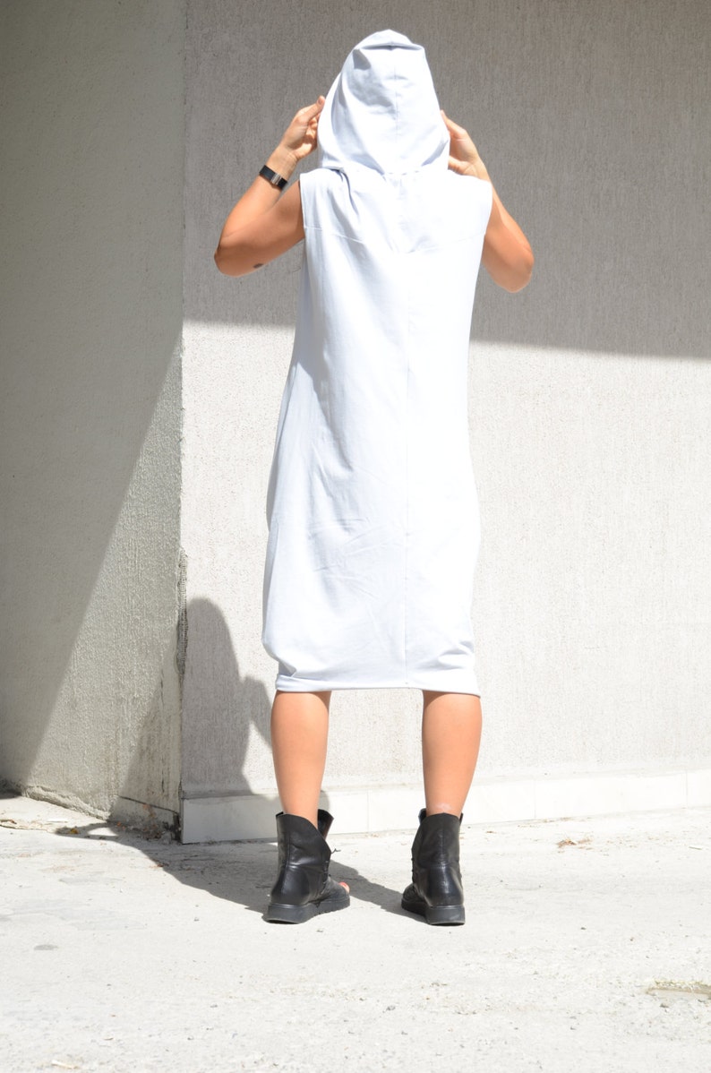 May include: A white sleeveless hooded dress with a long, flowing silhouette. The dress is made of a soft, comfortable fabric and has a relaxed fit. The model is wearing black ankle boots.
