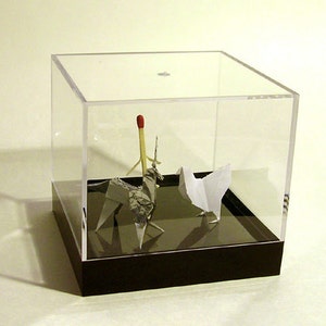 May include: A clear acrylic display case with a black base houses origami figures. A silver origami unicorn and a white origami bird stand near a matchstick. The case is on a light background.