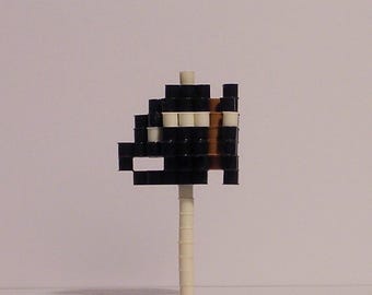 May include: A pixelated alien figure in black, white, and brown, mounted on a white stick. The alien has a blocky design, reminiscent of retro video games. The stick is cylindrical and appears to be made of plastic.