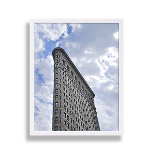 May include: A framed print of the Flatiron Building in New York City. The iconic skyscraper is gray and has a triangular shape. The sky is blue with fluffy white clouds. The print is in a white frame.