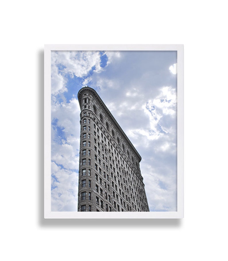 May include: A framed print of the Flatiron Building in New York City. The iconic skyscraper is gray and has a triangular shape. The sky is blue with fluffy white clouds. The print is in a white frame.