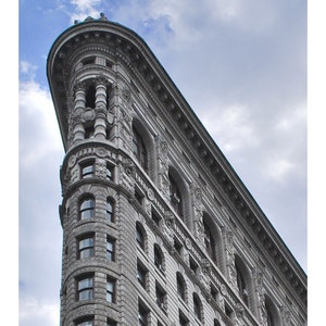 May include: Low-angle shot of the Flatiron Building in New York City. The building's unique triangular shape is visible, with ornate architectural details and numerous windows. The sky is overcast with clouds.