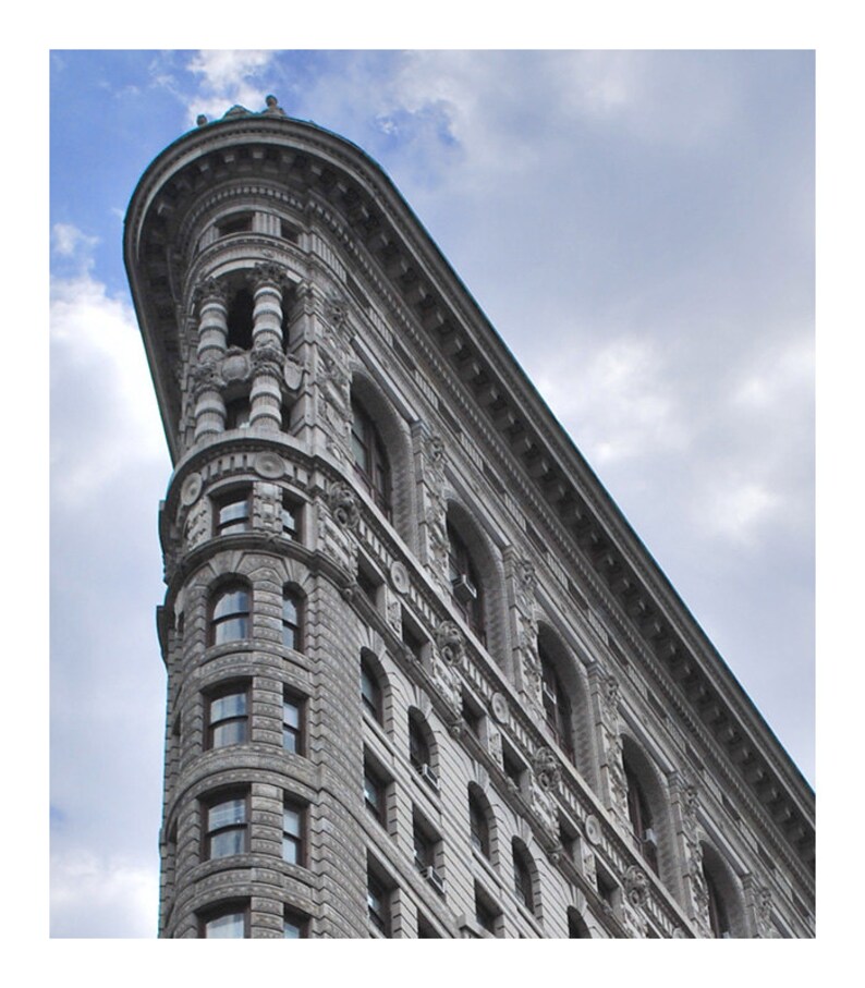 May include: Low-angle shot of the Flatiron Building in New York City. The building's unique triangular shape is visible, with ornate architectural details and numerous windows. The sky is overcast with clouds.