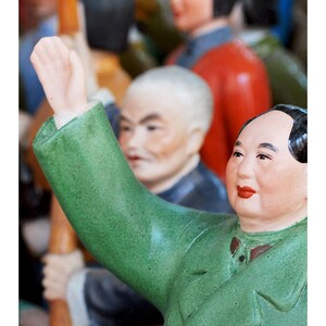 Puede incluir: Colecci&oacute;n de figuras de cer&aacute;mica, con una figura con chaqueta verde levantando el brazo. Las figuras tienen rasgos faciales detallados y est&aacute;n pintadas en varios colores. El fondo est&aacute; borroso, lo que sugiere un mercado o una exhibici&oacute;n.