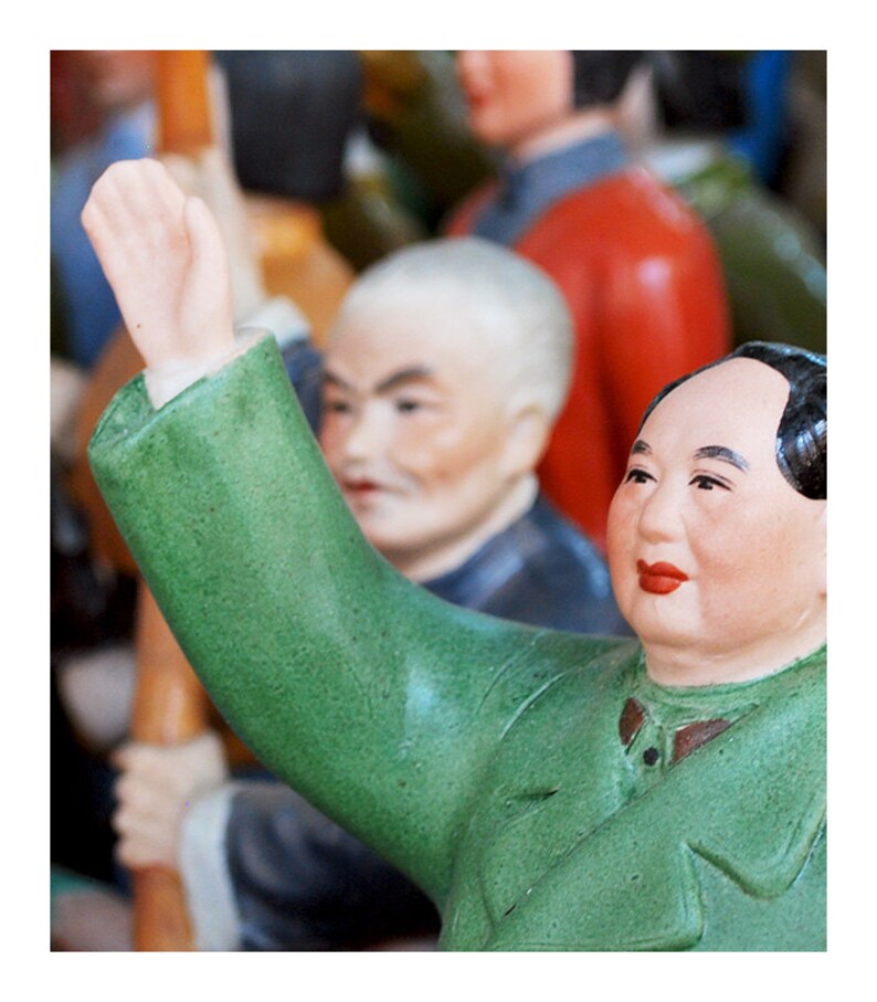 Puede incluir: Colecci&oacute;n de figuras de cer&aacute;mica, con una figura con chaqueta verde levantando el brazo. Las figuras tienen rasgos faciales detallados y est&aacute;n pintadas en varios colores. El fondo est&aacute; borroso, lo que sugiere un mercado o una exhibici&oacute;n.