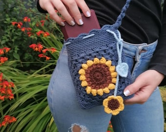 May include: A navy blue crochet crossbody bag with a sunflower design. The bag features a brown center, yellow petals, and a blue strap. A small yellow flower and a blue heart charm dangle from the bag.