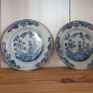 Pode incluir: Dois pratos de porcelana azul e branco com um design floral. Os pratos têm uma borda dourada e são decorados com flores azuis e brancas e uma pequena casa.