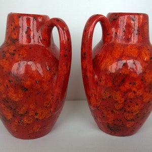 Puede incluir: Dos jarrones de cerámica roja con un esmalte moteado naranja y negro. Los jarrones tienen un cuerpo redondeado y un asa ancha y curva.