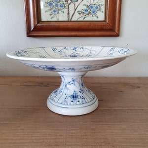 Puede incluir: Un soporte para tartas de cerámica blanca con un patrón floral azul y blanco. El soporte tiene una base redonda y un tallo alto y delgado.