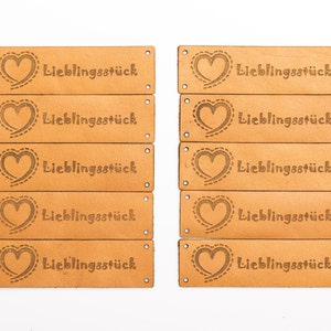 May include: A collection of rectangular, light brown leather tags. Each tag features a heart design with a dotted outline and the word "Lieblingsst&uuml;ck" in a serif font. The tags have small holes at each end.