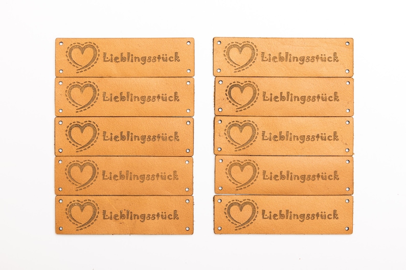 May include: A collection of rectangular, light brown leather tags. Each tag features a heart design with a dotted outline and the word "Lieblingsst&uuml;ck" in a serif font. The tags have small holes at each end.