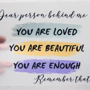 May include: A white card with the text "Dear person behind me, YOU ARE LOVED, YOU ARE BEAUTIFUL, YOU ARE ENOUGH, Remember that!" in black script and brushstroke accents of blue, yellow, and purple. The card is held by a hand.