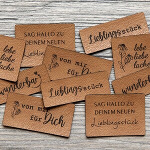 Könnte beinhalten: Eine Ansammlung brauner Lederetiketten mit verschiedenen Schriftzügen in schwarzer Schrift. Einige Etiketten zeigen florale Illustrationen. Sätze wie "lebe liebe lache" und "Lieblingsstück".