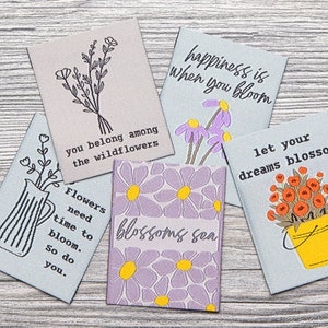 May include: Five cards with floral designs and inspirational quotes. The quotes are: "happiness is when you bloom", "you belong among the wildflowers", "Flowers need time to bloom. So do you.", "blossoms sen", and "let your dreams blossom".
