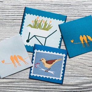 Op de afbeelding: Een verzameling vierkante stoffen patches met geborduurde ontwerpen. De patches tonen vogels op een draad, een kruiwagen met bloemen en een vogel op een tak. De patches zijn in blauwe en grijze tinten.