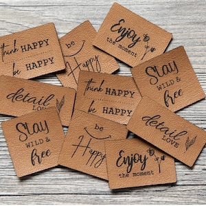 Puede incluir: Una colección de nueve etiquetas de cuero marrón con varias frases y diseños inspiradores, que incluyen "Think Happy, Be Happy", "Stay Wild & Free", "Detail Love" y "Enjoy the Moment".