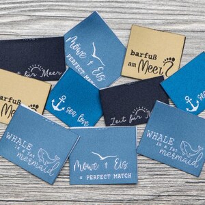 May include: A collection of rectangular fabric labels in shades of blue, navy, and tan. The labels feature various white or black printed designs including anchors, whales, seagulls, and text such as "sea love," "WHALE is a fat mermaid," and "barfuß am Meer."