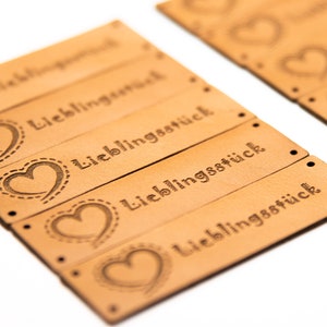May include: A collection of tan leather tags, each featuring the word "Lieblingsst&uuml;ck" and a heart motif. The tags are pre-punched with small holes for attachment. The heart design is outlined with a dotted stitch.
