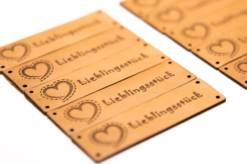May include: A collection of tan leather tags, each featuring the word "Lieblingsst&uuml;ck" and a heart motif. The tags are pre-punched with small holes for attachment. The heart design is outlined with a dotted stitch.