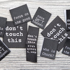 May include: A collection of black rectangular fabric labels with white text. The labels feature phrases including "don't touch this," "focus on the good," "make SH*T happen," and "who is Chuck?" in a sans-serif font, arranged in a scattered layout.