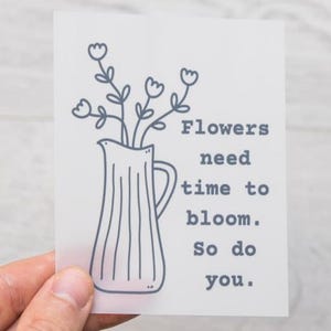 Könnte beinhalten: Eine weiße Karte mit einer Strichzeichnung einer Vase mit Blumen. Der Text lautet: "Blumen brauchen Zeit zum Blühen. Du auch."