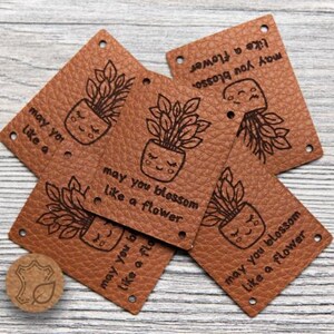May include: Brown leather tags with the text "may you blossom like a flower" and a potted plant illustration. Two round leather pieces with a leaf design are also visible. The tags have small holes for attaching.