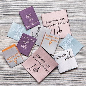Puede incluir: Una colección de etiquetas de tela de colores con varias frases alemanas impresas en ellas, incluyendo "Sag zu mir!" y "Staunen ist unfreiwilliges Lob".
