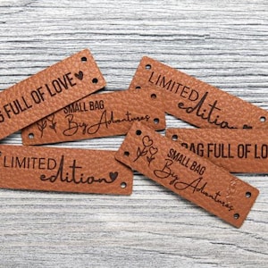 Könnte beinhalten: Mehrere braune Lederetiketten mit den eingravierten Aufschriften „BAG FULL OF LOVE“ und „LIMITED edition“. Die Etiketten haben kleine Löcher zum Annähen und dekorative Blumen- und Herzmotive.