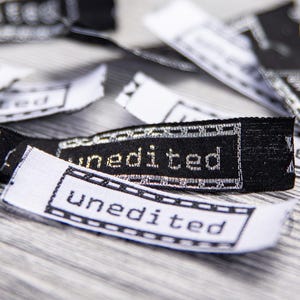 Könnte beinhalten: Nahaufnahme von mehreren schwarz-weißen Stoffetiketten mit dem Wort "unedited" darauf gestickt. Die Etiketten sind rechteckig mit dekorativem Rand und liegen auf einer hellgrauen Oberfläche verstreut.