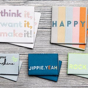 Könnte beinhalten: Eine Sammlung von bestickten Stoffaufnähern mit inspirierenden Sprüchen. Die Aufnäher zeigen Texte wie "think it, want it, make it!" und "HAPPY" in verschiedenen Farben. Andere Aufnäher sagen "create your own sunshine", "JIPPIE.YEAH" und "ROCK IT."