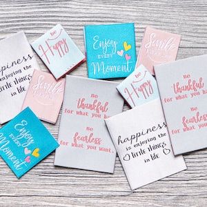 May include: Assorted fabric patches with embroidered inspirational quotes. The patches are in various colors, including light blue, pink, and gray. Quotes include "Enjoy Every Moment," "Be Happy," and "Happiness is enjoying the little things in life."
