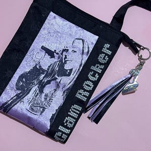 May include: A black and purple tote bag with a sequined design that says "Glam Rocker". The bag features a black and purple tassel and a silver charm. The bag has a black strap and a silver zipper.