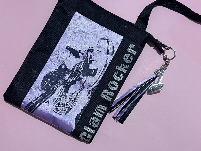 May include: A black and purple tote bag with a sequined design that says "Glam Rocker". The bag features a black and purple tassel and a silver charm. The bag has a black strap and a silver zipper.