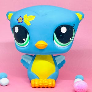 May include: A blue and yellow bird-shaped toy with large, expressive eyes. The toy has a flower and leaf design on its head and orange feet. The background is a solid pink color. The toy is a collectible item.