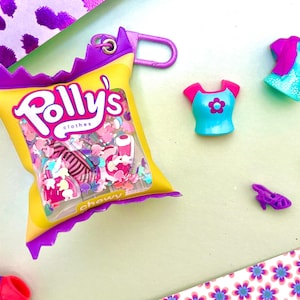 May include: A yellow and purple Polly's clothes snack bag keychain filled with miniature doll accessories, including a blue and pink shirt, a purple and blue top, a single purple high-heeled shoe, and a tiny yellow accessory piece.