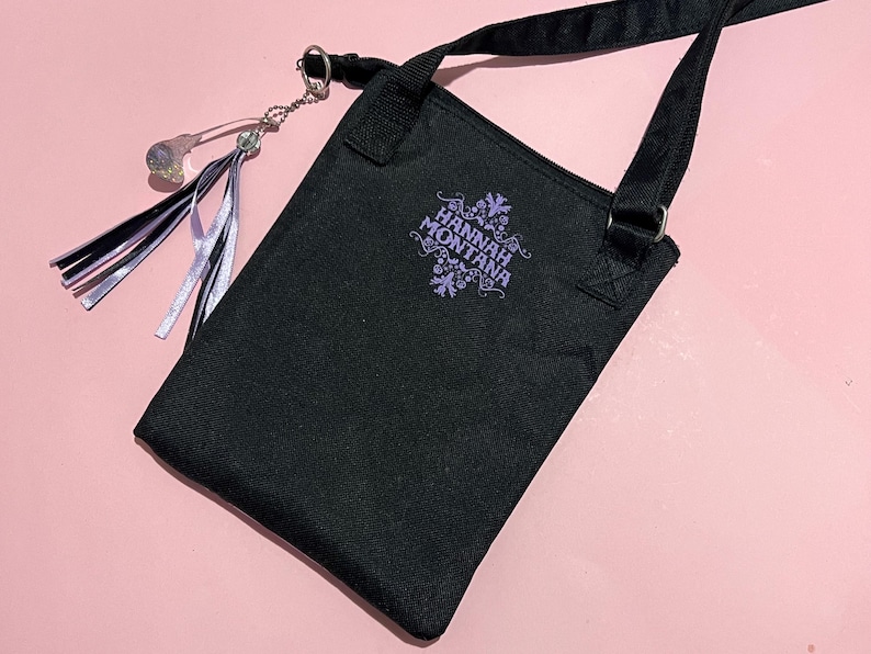 May include: Black canvas shoulder bag with a purple embroidered logo that says "Hannah Montana". The bag has a silver chain with a crystal charm attached.