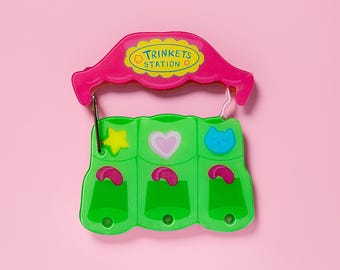 Puede incluir: Un expositor de baratijas rosa y verde con el texto "TRINKETS STATION". El expositor tiene tres compartimentos verdes con un diseño de estrella, corazón y gato. El expositor está colgado sobre un fondo rosa.