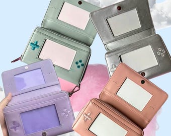 May include: Four rectangular zippered wallets designed to resemble handheld gaming consoles. The wallets come in pastel purple, mint green, metallic silver, and dusty rose, each featuring a central clear window and embossed directional pad buttons.
