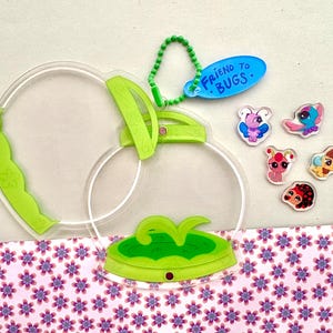 Peut inclure: Un attrape-insectes en plastique transparent avec une poignée verte et une base verte. L'attrape-insectes est doté d'un charme vert "Ami des insectes" qui y est attaché. Il y a aussi plusieurs petites figurines d'insectes en plastique sur une surface à motifs rose et blanc.