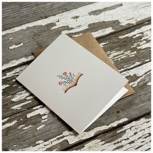 May include: A blank greeting card with a watercolor illustration of an open book with pink flowers growing from the pages.