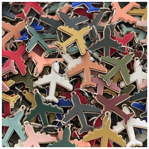 May include: A collection of airplane-shaped keychains in various colors, including red, white, yellow, green, and blue. Each keychain has a silver ring for attaching keys. The keychains are made of a smooth material and are scattered across the image.